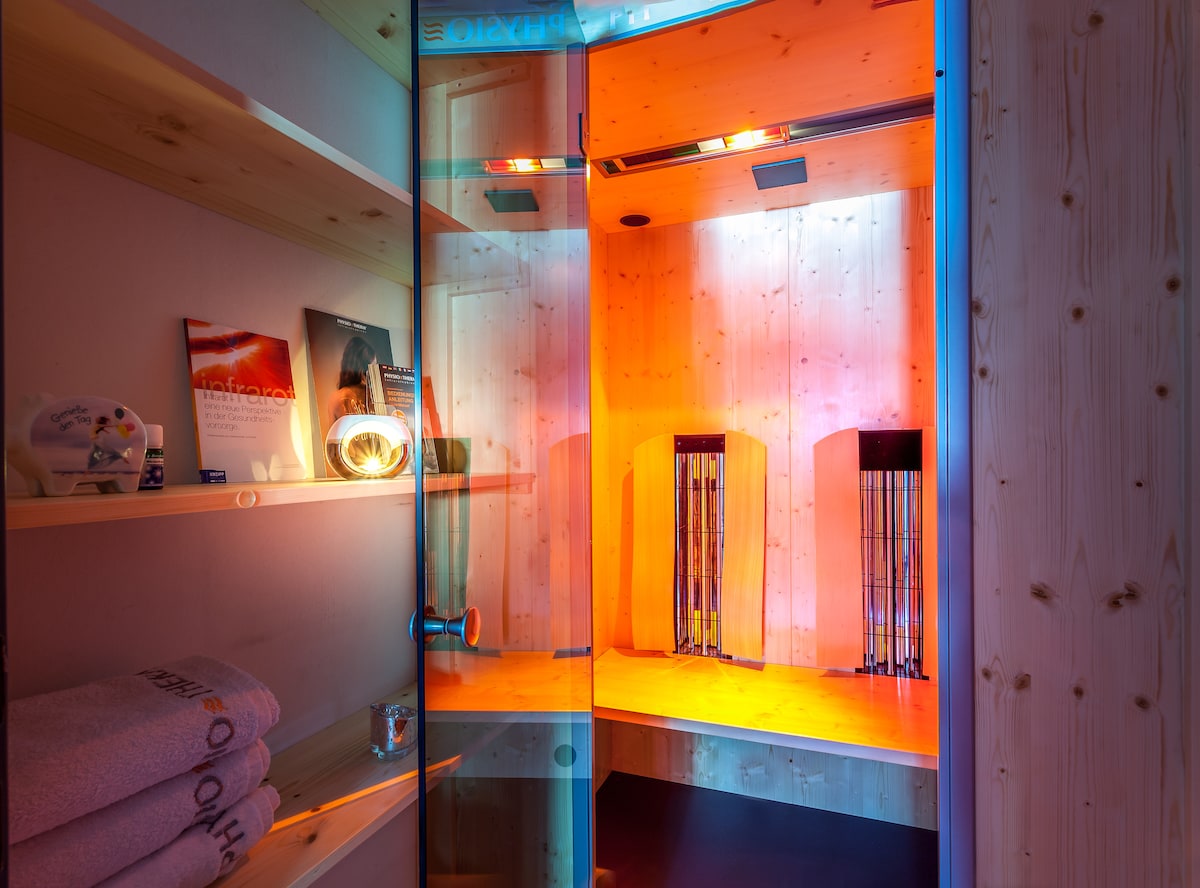 A modern sauna featuring wooden paneling and ambient lighting. The glass door allows for a view of the spacious interior, where wooden benches are illuminated with soft hues of orange and blue. Towels are neatly folded on a shelf nearby.