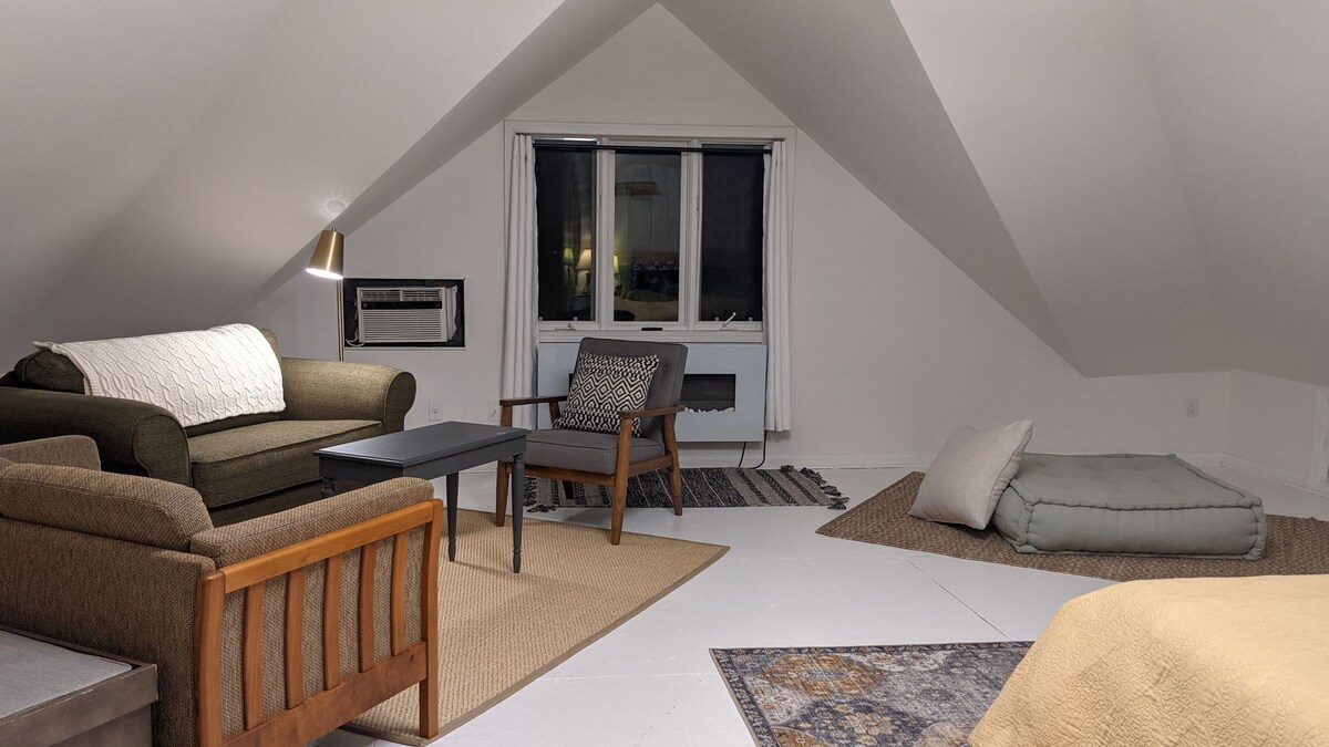 A spacious sitting area is featured under a sloped ceiling, furnished with a sofa, an accent chair, and a small coffee table. Natural light enters through a window with light curtains, and a soft area rug adds warmth to the floor.