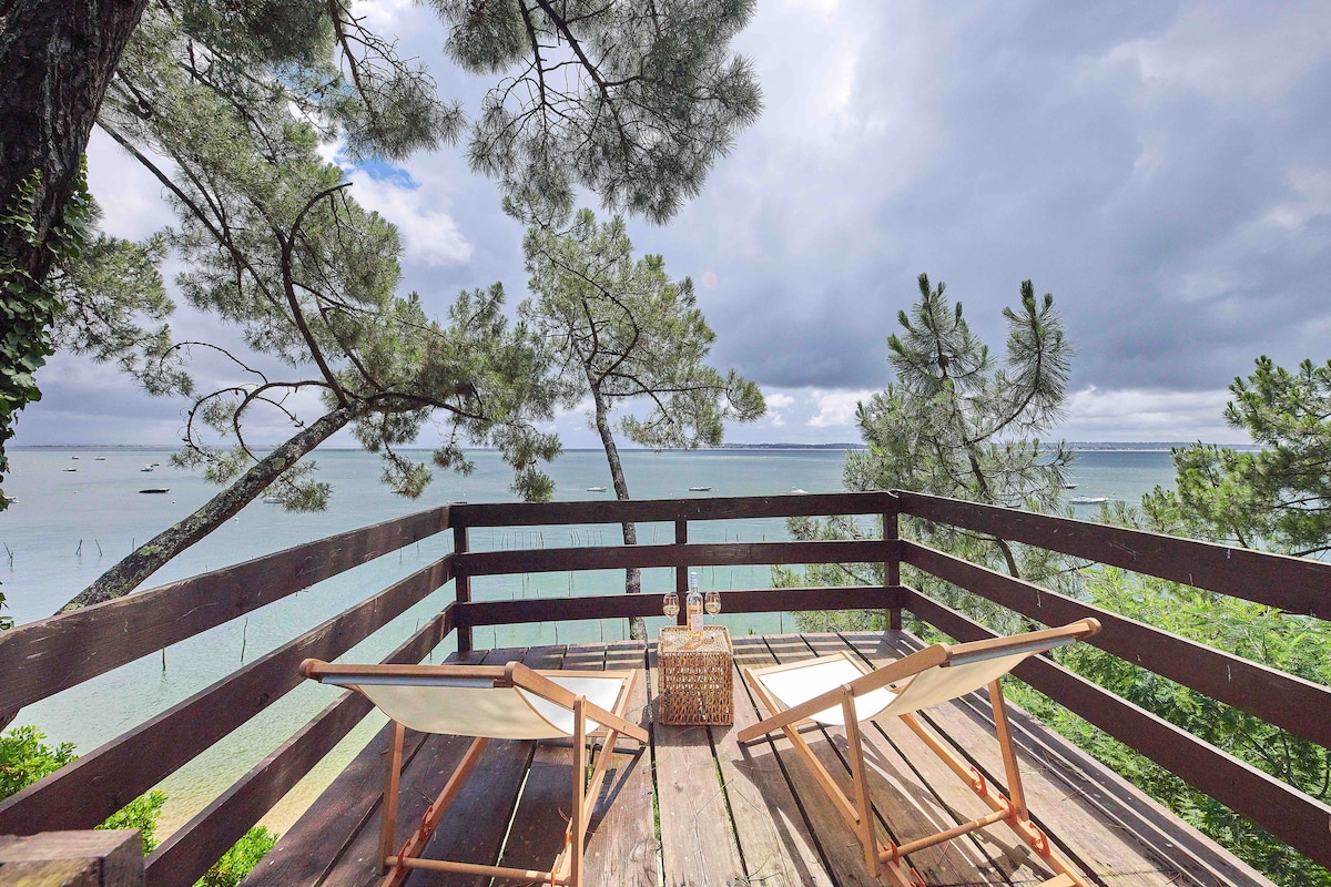 A furnished wooden deck offers a serene view of the water, surrounded by tall trees. Two lounge chairs are positioned invitingly, with a small table nearby. The sky above is partly cloudy, enhancing the peaceful environment.