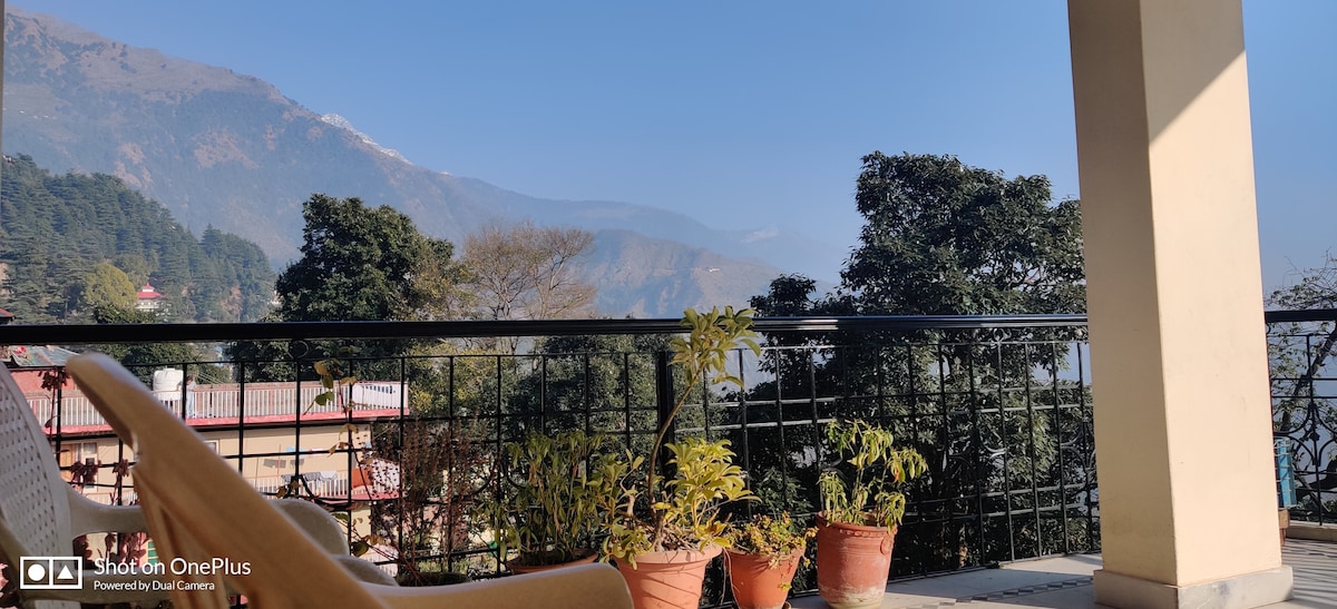 A balcony view showcases surrounding hills under a clear sky. Potted plants add greenery, while the railing provides a sense of openness. The distant mountains provide a serene backdrop, enhancing the tranquil outdoor space.