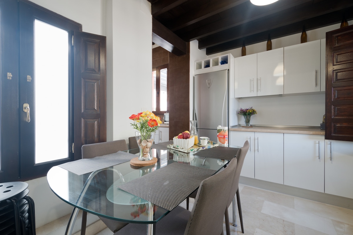A modern kitchen and dining area is showcased, featuring a glass dining table surrounded by four upholstered chairs. White cabinetry is complemented by wooden touches, and fresh flowers adorn the table. Natural light enters through large windows, enhancing the warm tones of the space.