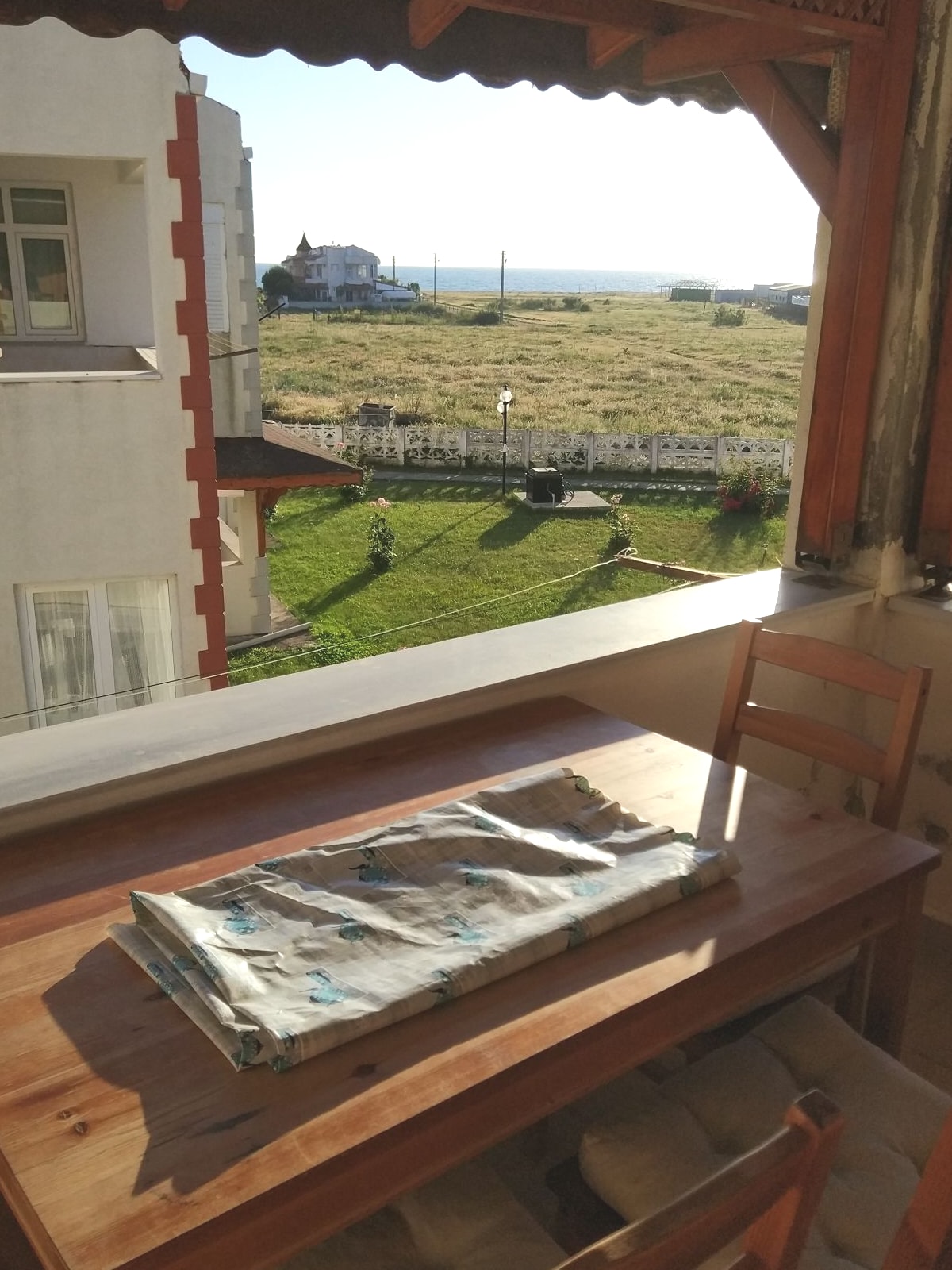A wooden dining table is positioned on the balcony, accompanied by a chair. The table is adorned with a folded cloth featuring a subtle pattern. The view beyond reveals a grassy landscape and distant sea, offering a sense of openness and natural light.