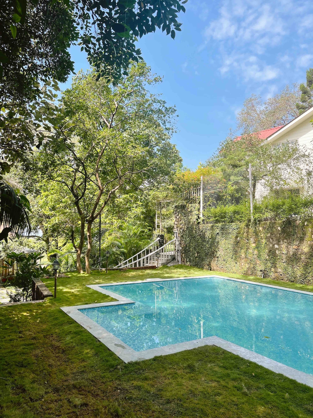 A serene swimming pool is surrounded by lush greenery and trees, with steps leading to a higher level. The clear water reflects the blue sky, inviting relaxation. A well-maintained lawn frames the pool area, creating a peaceful escape in the outdoors.