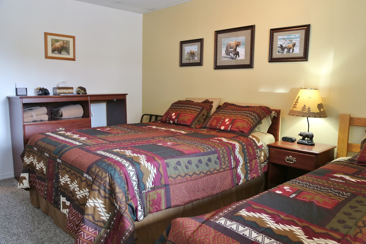 A wildlife-themed bedroom is displayed, featuring a spacious queen bed and a twin bed topped with patterned bedding. Framed wildlife photos adorn the walls, while a bedside lamp provides soft illumination. A dresser with neatly stacked towels is present, enhancing functionality.
