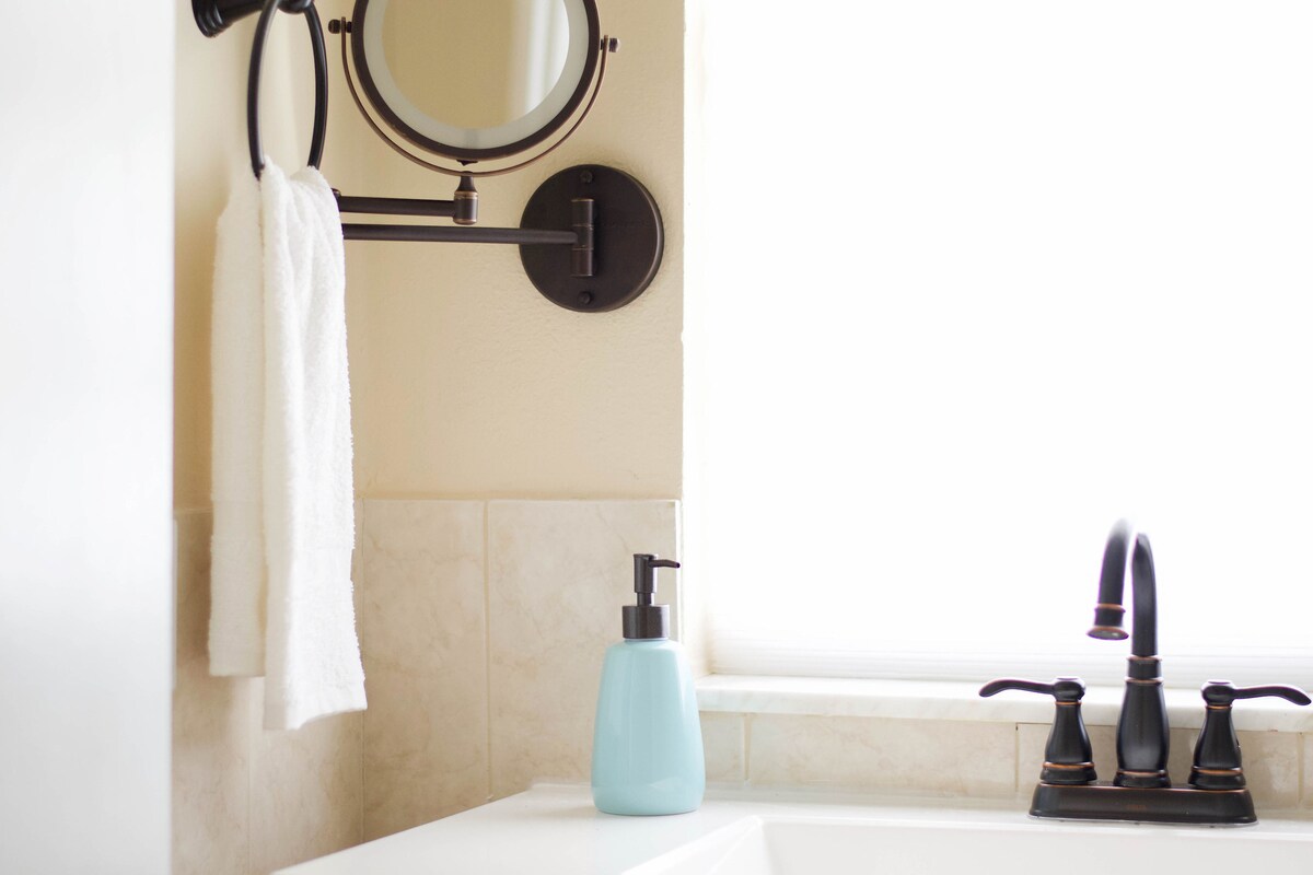 A well-appointed bathroom corner features a stylish wall-mounted round mirror adjacent to a towel rack holding a white towel. A light-colored soap dispenser rests on the counter, and elegant dark fixtures are visible near the window, ensuring natural light enhances the space.