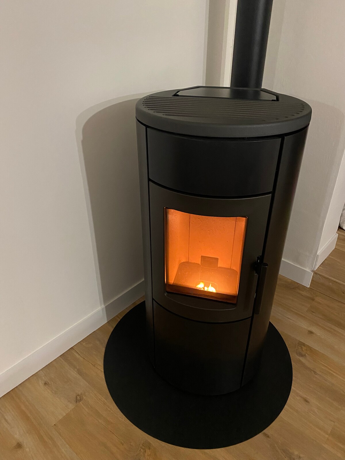 A modern black stove is positioned against a white wall. The round base is matched by a circular platform beneath it. A warm glow emanates from the window, hinting at a cozy fire inside, creating a welcoming focal point in the room.