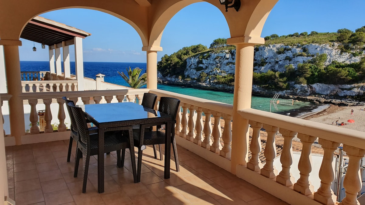 A shaded terrace features a sturdy dining table surrounded by four chairs, offering scenic views of the coastline. The ocean sparkles under the sun, and a sandy beach is visible nearby, framed by natural greenery and rocky cliffs.