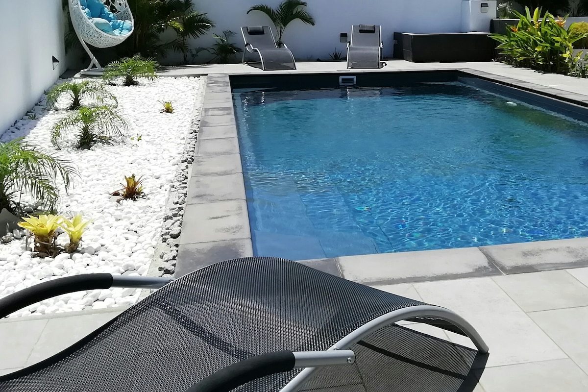 The salt-water pool is surrounded by a neat patio and features modern lounge chairs for relaxation. Lush greenery and white decorative stones create a serene atmosphere, enhancing the outdoor space.
