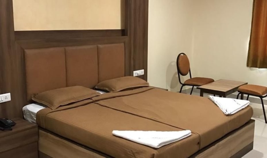 A spacious room features a large, neatly made bed with brown bedding and two pillows. A small wooden table and two chairs occupy one corner, while a telephone is placed on the bedside table. Natural light enters through a window covered with light curtains.