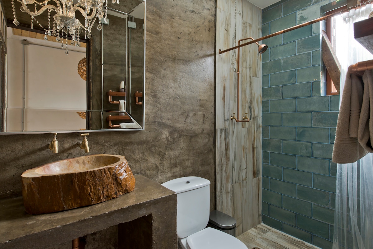 A modern bathroom features a unique stone sink set into a concrete vanity. The shower area is designed with turquoise tiles, complemented by copper fixtures. A large mirror reflects the elegant chandelier above. Natural light filters through a sheer curtain.