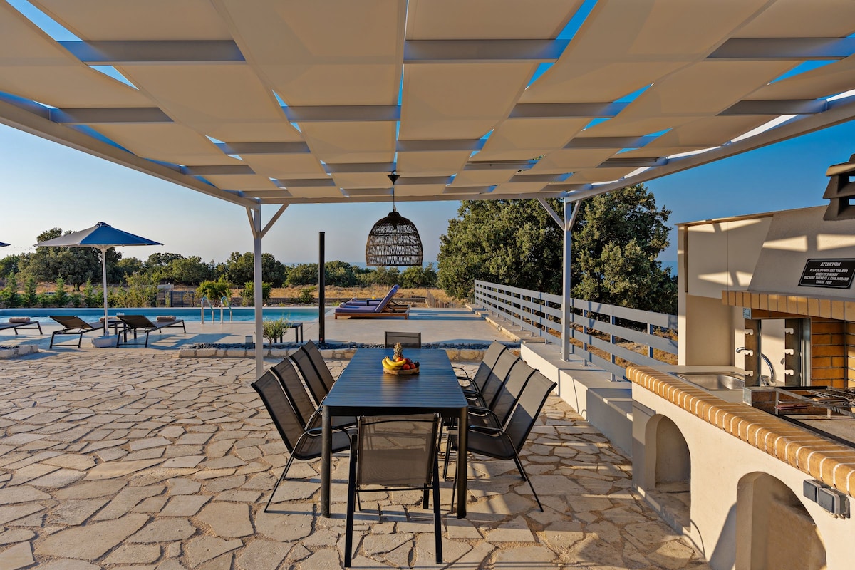 A shaded dining area features a long table surrounded by black chairs, overlooking a serene setting. An outdoor barbecue area is visible nearby, with a backdrop of the pool and lush greenery enhancing the tranquil atmosphere.