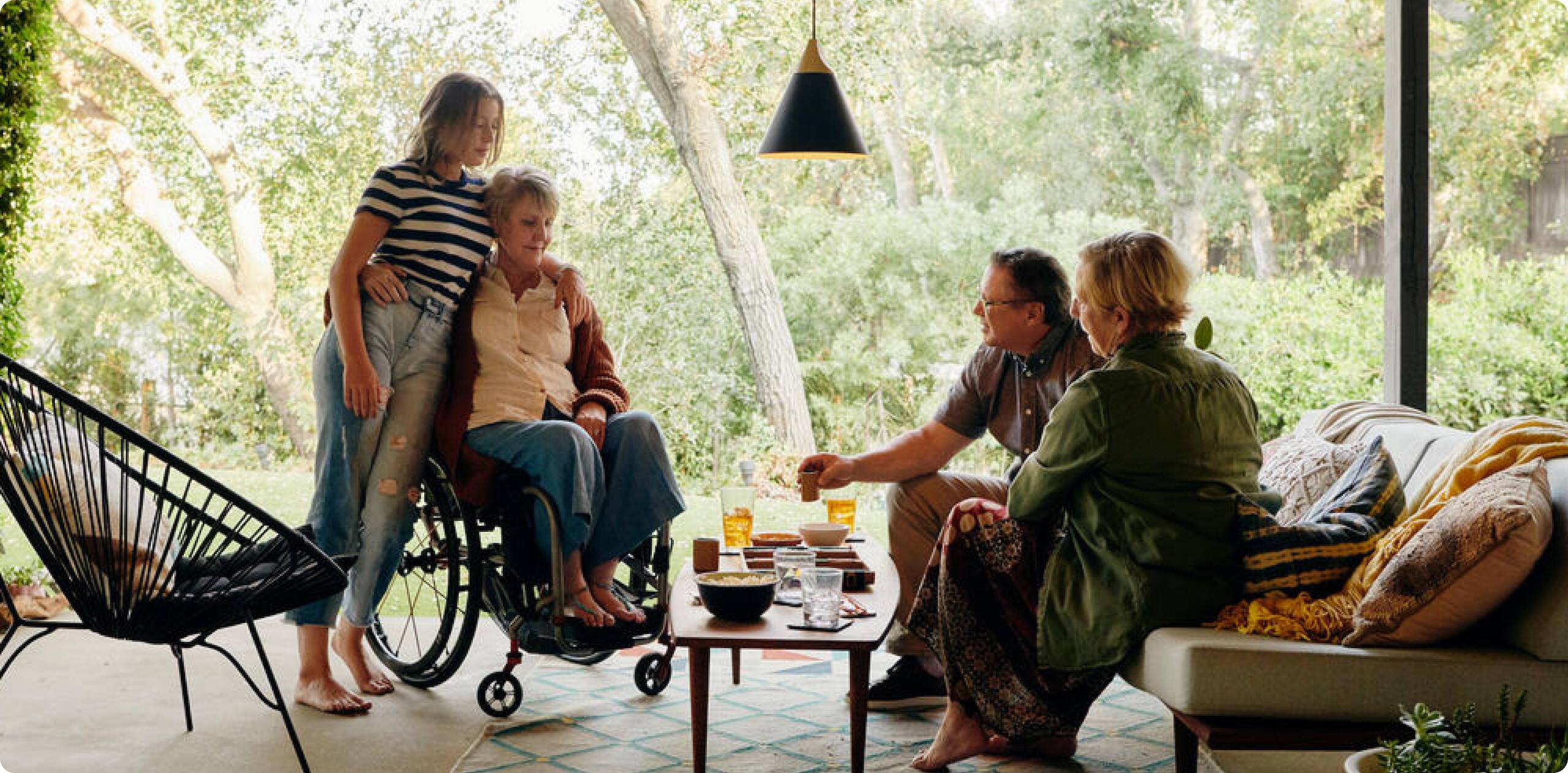 Tres generaciones de familias riendo en un alojamiento accesible en Airbnb. Enfrente del familiar mayor se encuentra una silla de ruedas. Tres generaciones de familias riendo en un alojamiento accesible en Airbnb. Enfrente del familiar mayor se encuentra una silla de ruedas.