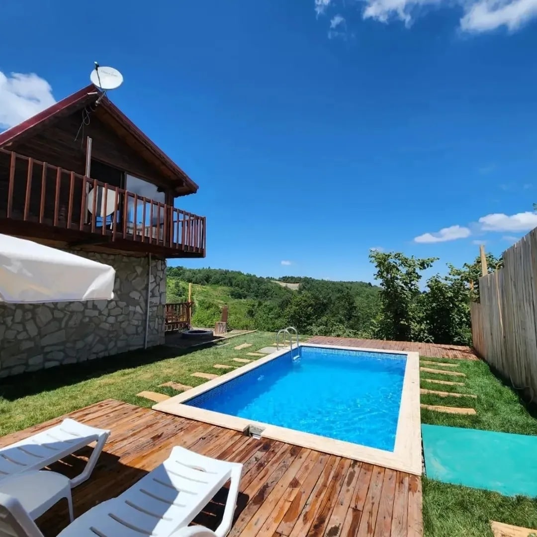 A clear blue sky is visible above a refreshing pool framed by green grass and wooden decking. A charming two-story house is positioned nearby, with a balcony overlooking the scenic landscape. Lounge chairs are placed beside the pool for relaxation.