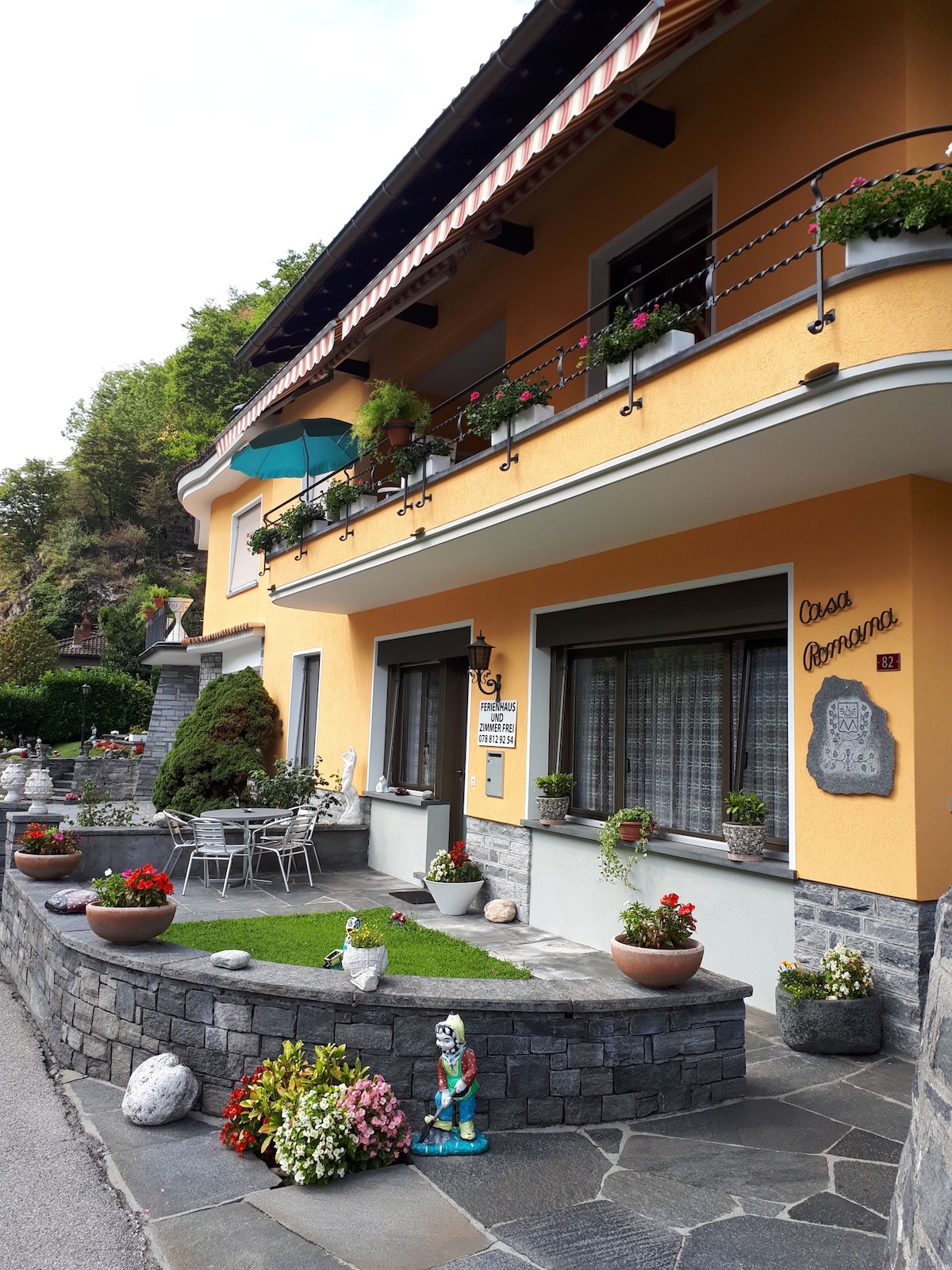 A charming two-story house features a bright yellow exterior and a welcoming porch area. Potted plants adorn the windows, and a small table with chairs is set on the patio. Decorative stones and flowers create a pleasant garden atmosphere.