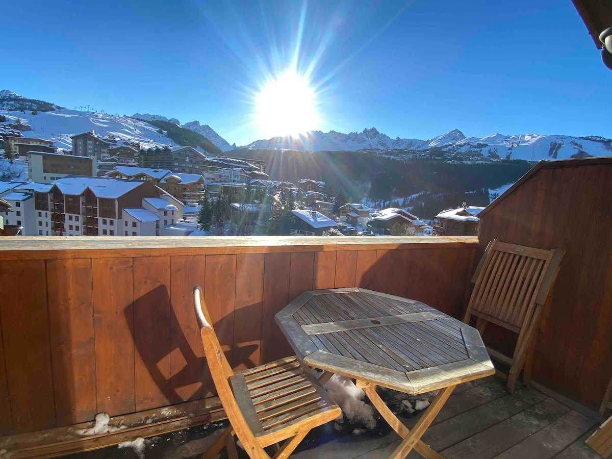 A wooden terrace features a round table with two matching chairs, overlooking a scenic mountain view. The morning sun shines brightly over snow-covered peaks, creating a picturesque backdrop for outdoor relaxation.