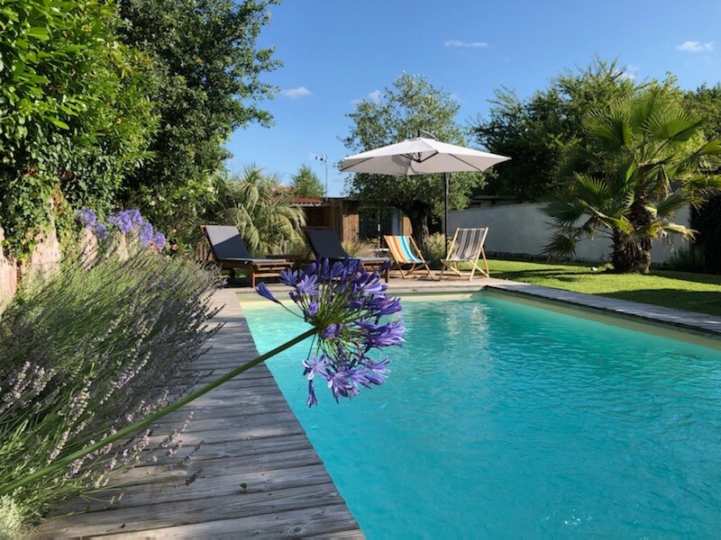 A serene outdoor space is showcased, featuring a clear swimming pool surrounded by wooden decking and lounge chairs. Lush greenery, including palm trees and lavender, complements the setting, while a large umbrella provides shade for relaxation. A bright blue sky adds to the inviting atmosphere.