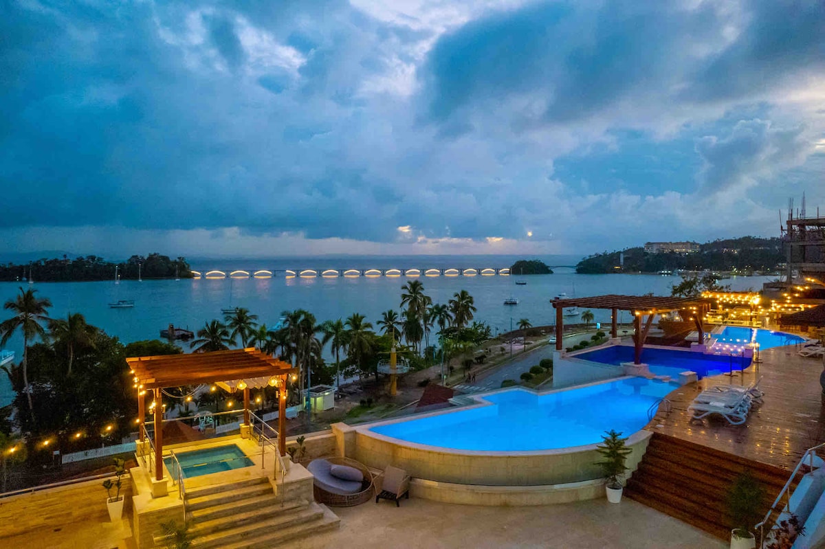 A scenic view of the pool area showcases two blue pools and a jacuzzi, surrounded by lounge chairs and palm trees. In the background, Samaná Bay glistens under a cloudy sky, reflecting hues of blue and grey. Soft lights illuminate the outdoor space, enhancing the relaxing atmosphere.