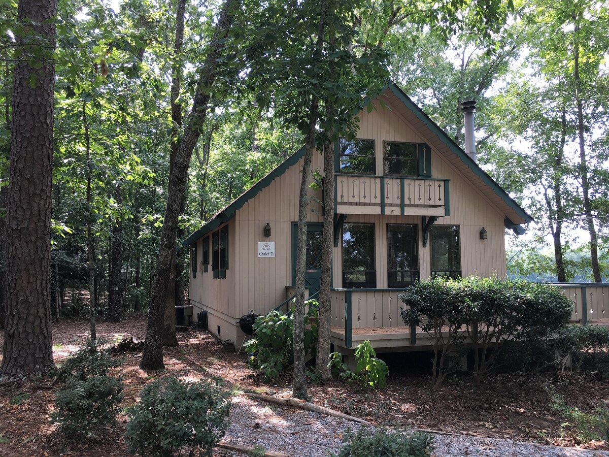 A charming cabin exterior, nestled among tall trees. The wooden structure features large windows, allowing natural light to fill the interior. A welcoming porch extends from the front, framed by neatly trimmed shrubs and a gravel path, creating a serene atmosphere.