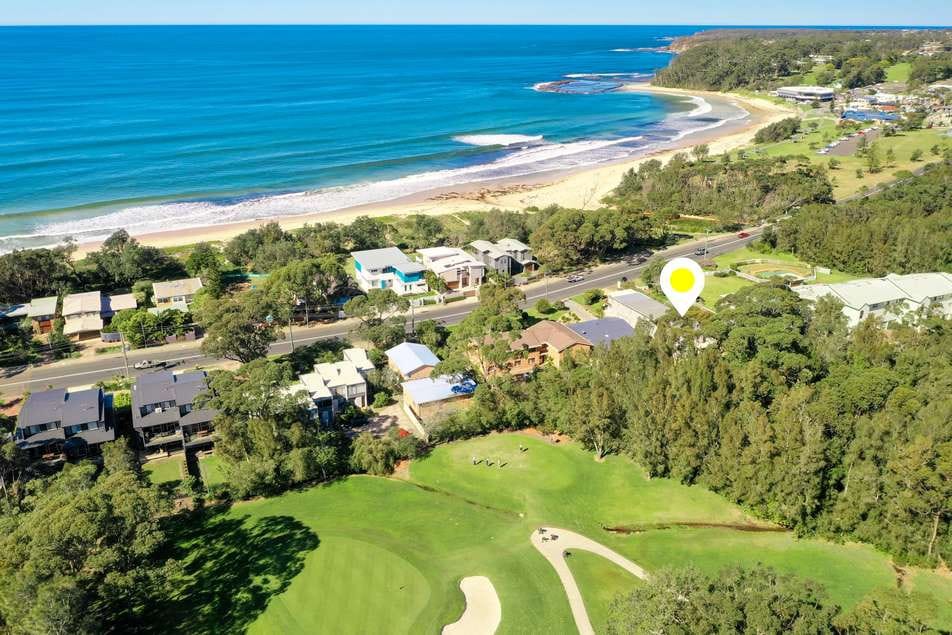 An aerial view captures the coastal landscape, featuring the ocean's gentle waves lapping against the beach. The lush green golf course can be seen in the foreground, with residential houses lining the shore and trees dotting the area, creating a serene beachfront setting.