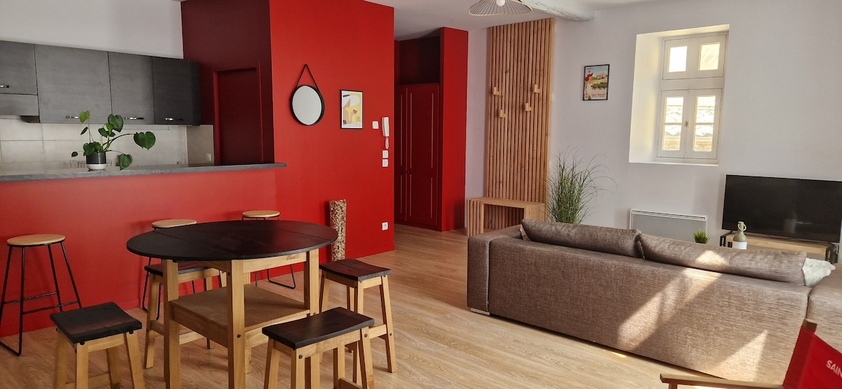 A bright living area showcases a red accent wall, complemented by a modern kitchenette in the background. A round dining table is surrounded by wooden stools, while a spacious sofa sits in front of a television. Natural light filters through the window.