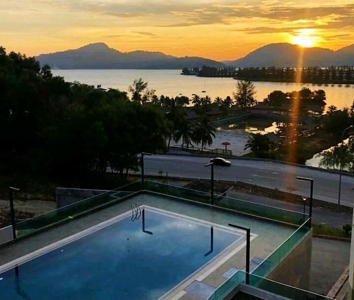 A serene view of a swimming pool overlooking a river and mountains, with the sun setting on the horizon. Soft sunlight reflects off the pool's surface, enhancing the tranquil ambiance surrounded by greenery.