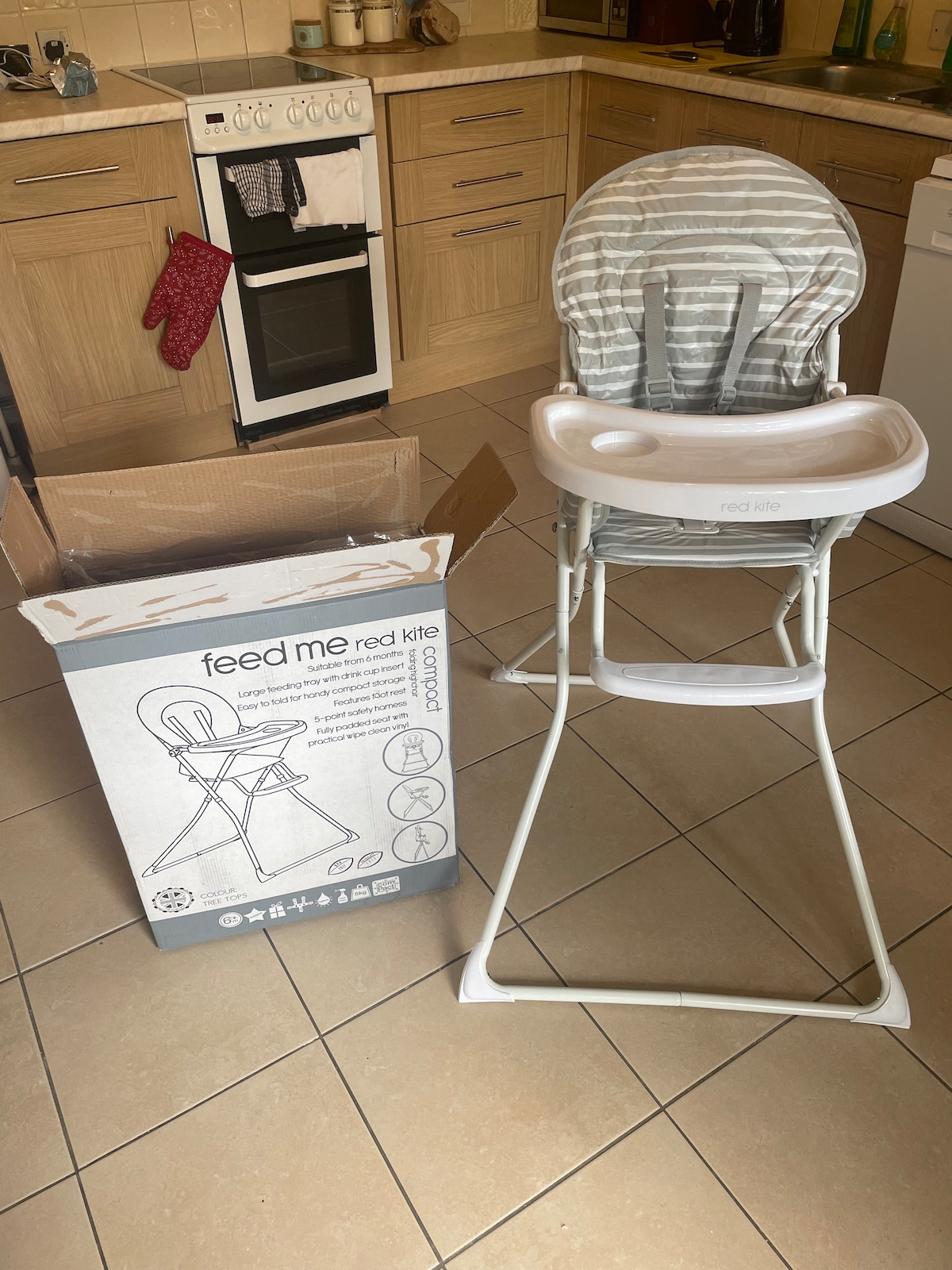 A high chair is positioned in a kitchen setting, featuring a gray striped seat and a white, molded tray. A partially opened cardboard box is nearby, displaying product information. The cabinetry in the background includes wooden finishes and various kitchen items.