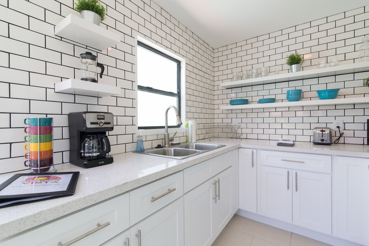 A modern kitchen is highlighted by white cabinetry and a gray countertop. Open shelving displays vibrant dishware, while a coffee maker and toaster are visible on the counter. Natural light streams through a window, enhancing the tiled backsplash design.