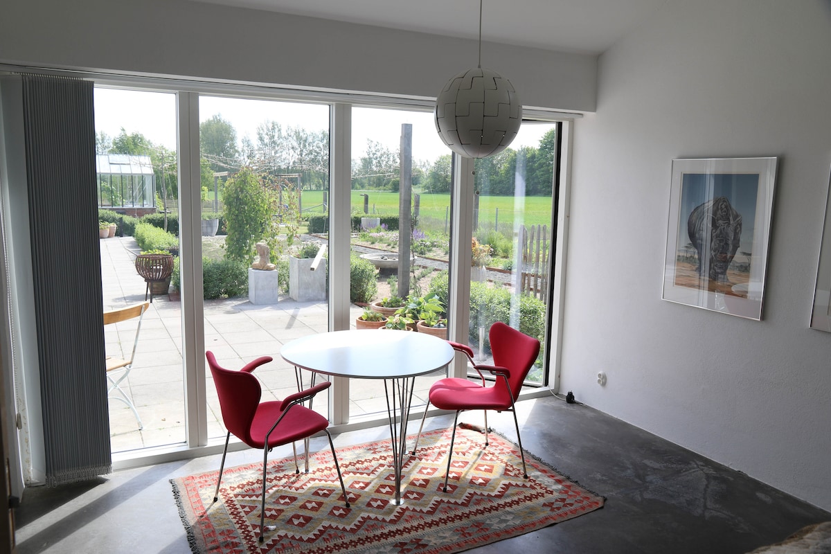 A well-lit dining area features a round table surrounded by two red chairs. Large glass doors provide an unobstructed view of the greenery outside, while a colorful rug lies beneath the table. A pendant light hangs from the ceiling, offering ambient illumination.