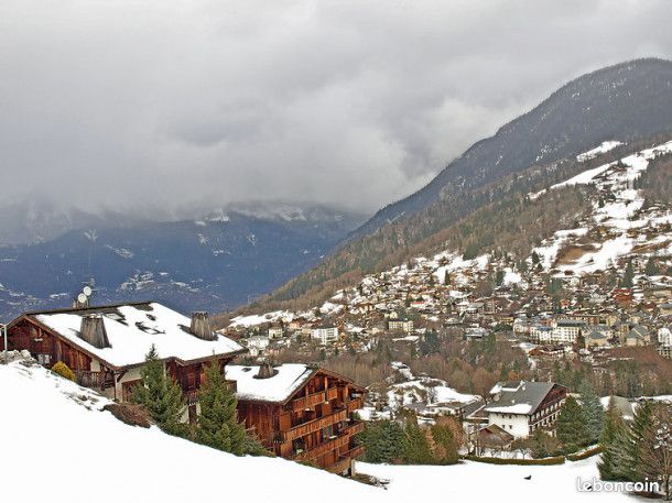 A scenic view of a mountainous landscape features snow-covered rooftops of chalets and the surrounding altitude. Dense trees intersperse the lower slopes, and a village is visible in the distance, framed by cloudy skies. The image conveys a tranquil winter atmosphere.