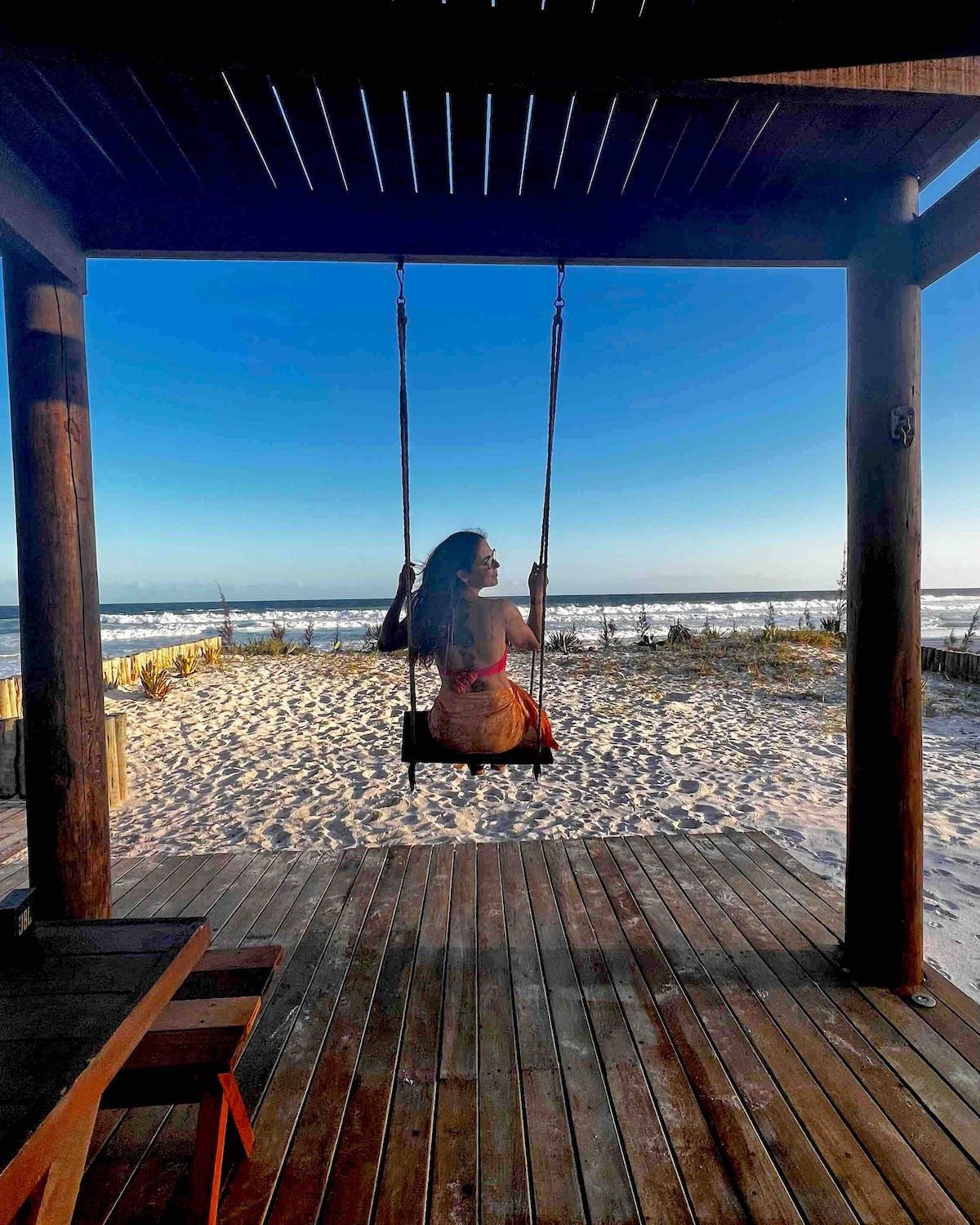 A swing hangs from a wooden structure, facing a sandy beach and the ocean. Gentle waves can be seen in the distance under a clear blue sky, creating a calm and tranquil environment. Wooden decking provides a rustic flooring, adding to the outdoor setting.