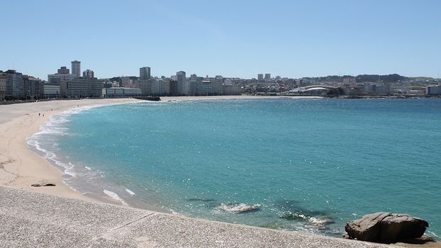 A sandy beach curves along a clear, turquoise ocean. The shoreline is dotted with distant buildings and stretches of vibrant coastline, while gentle waves lap against the shore. The scene is set under a bright, cloudless sky, capturing a serene coastal atmosphere.