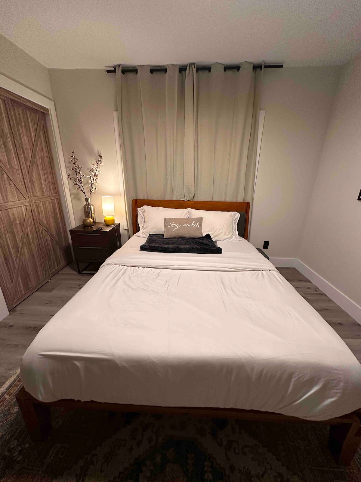 A serene bedroom features a wooden bed with a neatly made white bedspread and a dark throw placed at the foot. Soft curtains frame the window, while a bedside table holds a lamp and decorative elements, contributing to the peaceful ambiance.