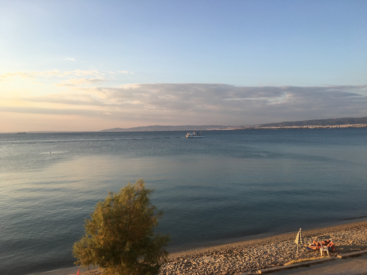 A serene view of the sea at sunset, displaying calm waters reflecting soft hues of orange and blue. A small boat can be seen in the distance, while a sandy beach with a single umbrella occupies the foreground, offering a peaceful coastal atmosphere.