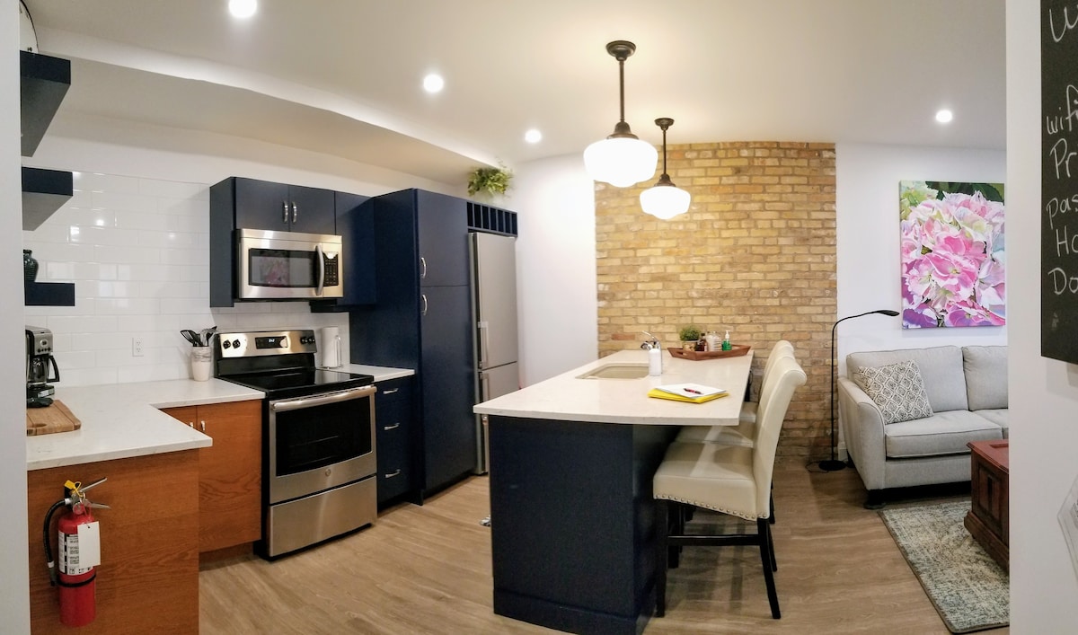 A modern kitchen area is visible, featuring dark blue cabinetry and stainless steel appliances. A white island separates the kitchen from a cozy living area, which includes a light grey sofa. Natural light floods the space, highlighting the exposed brick wall and a colorful floral art piece.