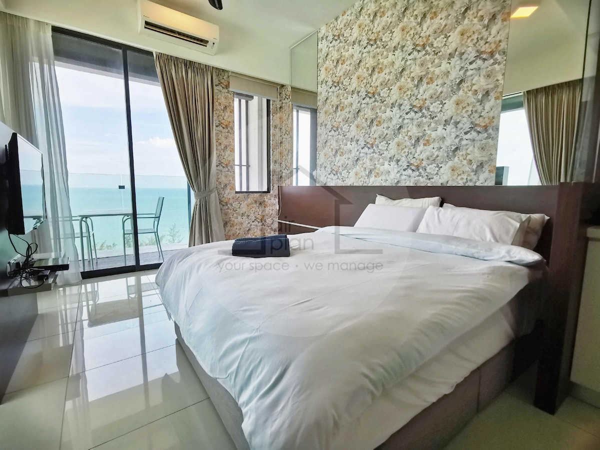 A cozy bedroom features a queen-size bed with white linens, large windows providing views of the ocean, and sheer curtains allowing natural light. A flat-screen TV is mounted on the wall opposite the bed. A small balcony is visible, enhancing the room's airy atmosphere.