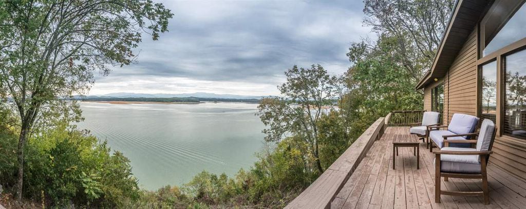 A panoramic view of Douglas Lake is visible from the spacious back deck. Comfortable seating is arranged on the wooden deck, surrounded by lush greenery. The calm water reflects the muted colors of the sky, creating a serene atmosphere overlooking the Smoky Mountains in the distance.
