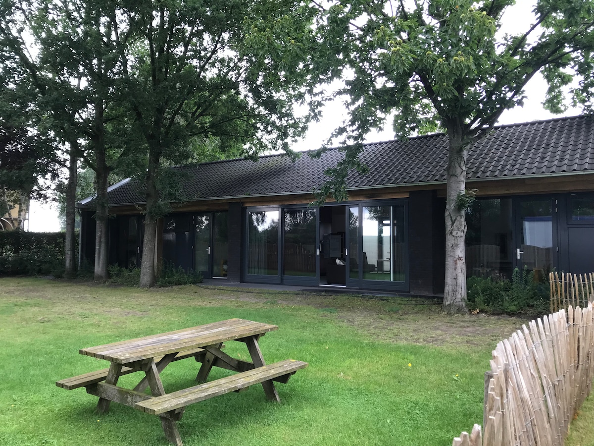 The exterior of the accommodation is visible, featuring a spacious lawn bordered by trees. A wooden picnic table sits on the grass, offering an outdoor seating area. Large glass doors provide an open view into the interior, enhancing the connection to the natural surroundings.