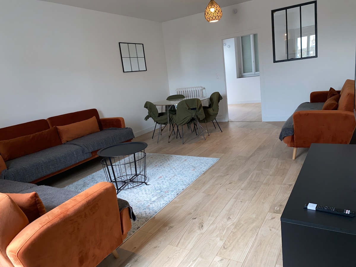 A spacious living area is furnished with two sofas in warm tones, complemented by a round dining table surrounded by green chairs. Natural light fills the space through large windows, enhancing the inviting atmosphere. A subtle area rug rests beneath a modern coffee table.