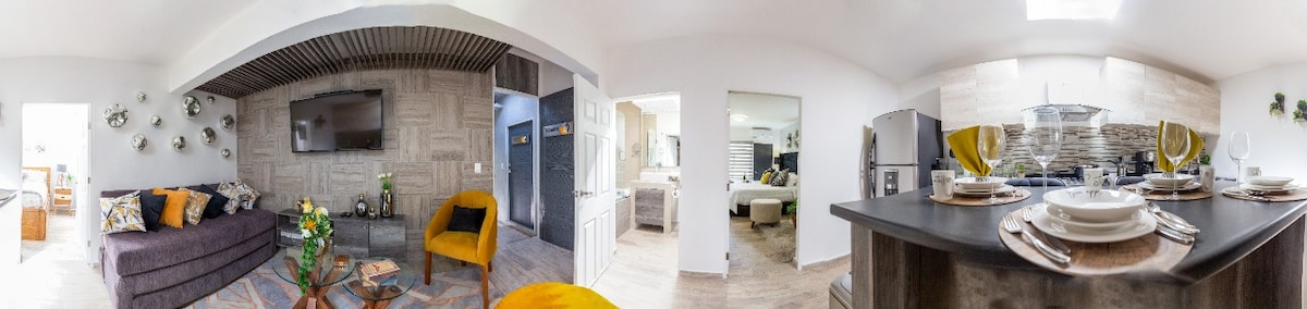 An open-plan living space is depicted, featuring a cozy seating area with a dark sofa and decorative pillows. A dining table is set for meals, with plates and glasses arranged neatly. The kitchen area shows modern appliances, enhancing the functional design of the suite.
