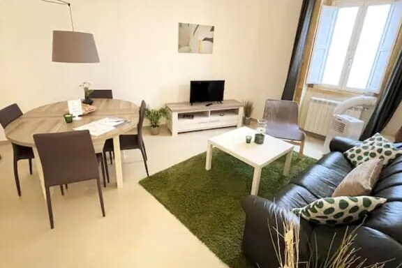 A bright living area features a round dining table surrounded by chairs, a cozy sofa adorned with patterned cushions, and a flat-screen television on a media console. Light enters through large windows, and green carpet adds a touch of color to the space.