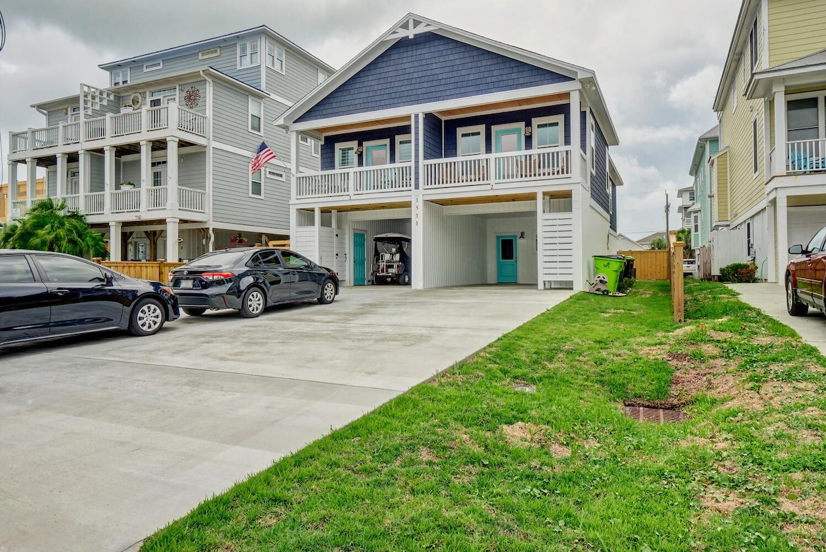 A modern townhome with a blue exterior and white accents is depicted. A spacious driveway accommodates multiple vehicles, with one covered parking spot visible. Nearby, green grass adds a touch of nature, while neighboring homes showcase varying architectural styles.