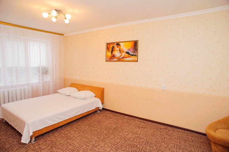 A spacious room features a light-colored wall and a soft carpet. A comfortable bed with two pillows is positioned against an orange upholstered base, while a piece of artwork is displayed above the bed. Natural light filters through a sheer-curtained window.