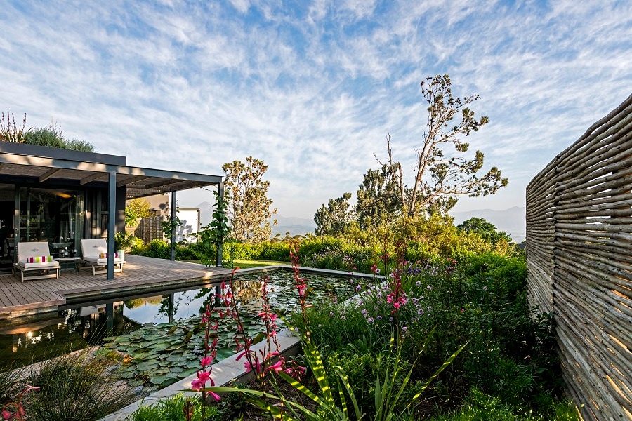 A serene outdoor space features a nature pool surrounded by lush greenery and flowering plants. Comfortable loungers are positioned on a wooden deck, offering views of the clear sky and distant mountains. The area is designed to blend harmoniously with its natural surroundings.