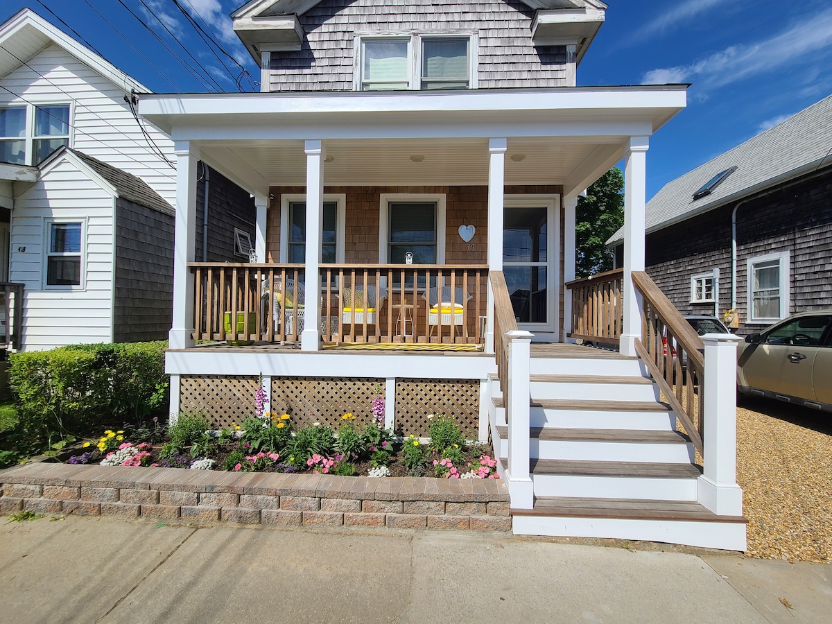 A welcoming front porch is featured, with two yellow chairs positioned beside a decorative heart. The light brown brick steps lead up to the entrance, flanked by a colorful flower bed with a variety of blooming plants. Clear blue skies complement the setting.