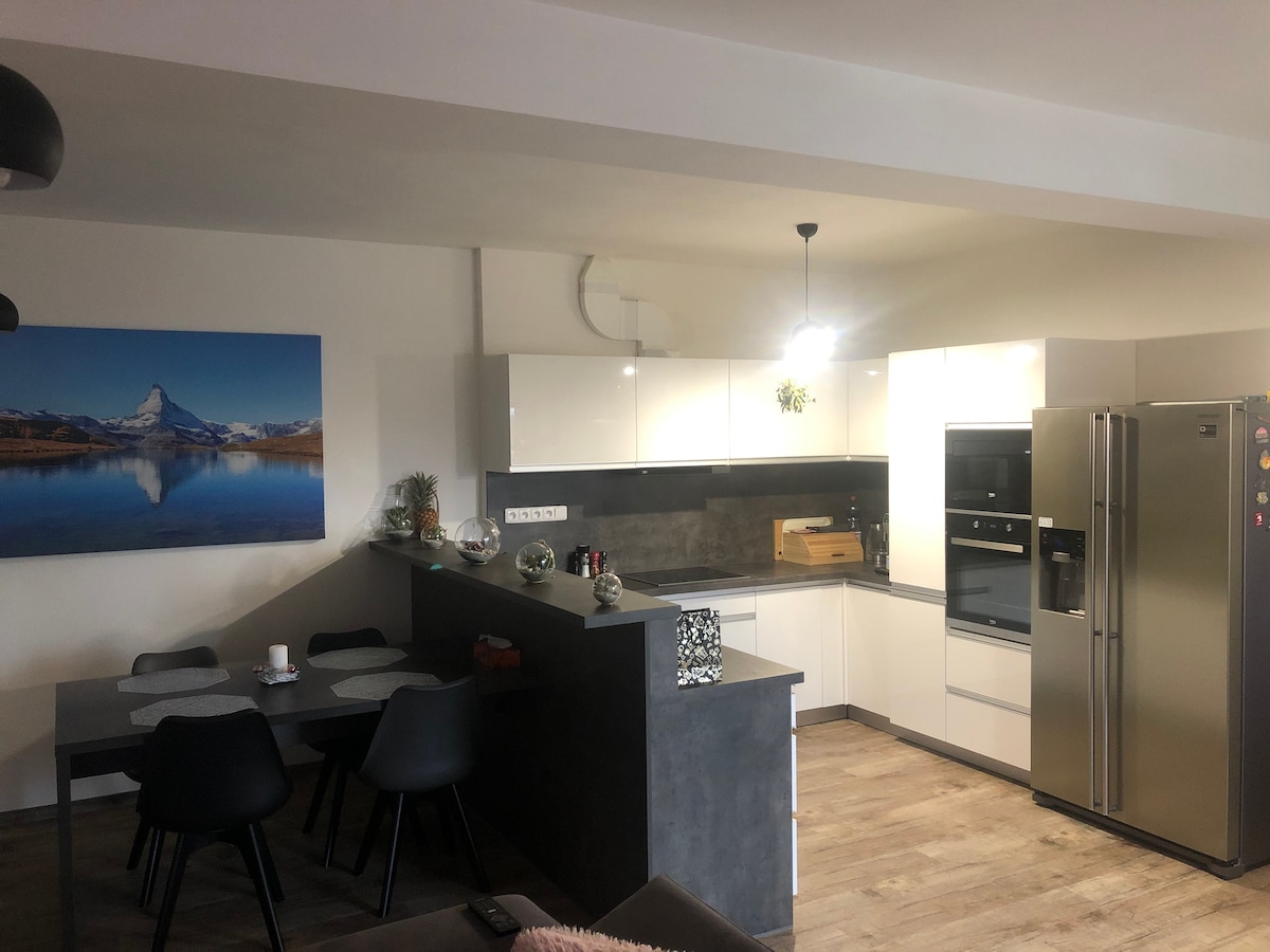 The modern kitchen is equipped with sleek white cabinetry and stainless steel appliances, including a large refrigerator. A dining table with black chairs accommodates guests, while a scenic mountain landscape painting adds a touch of nature to the space. Warm light illuminates the area.