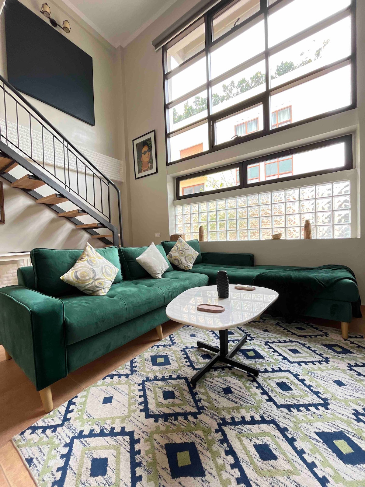 A modern living area features a green sofa adorned with decorative pillows. A coffee table sits in front amid a patterned area rug. Large windows allow natural light to fill the space, and a staircase leads to an upper level.