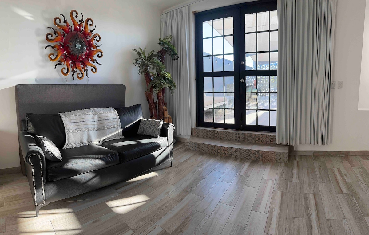 A spacious living area features a dark sofa complemented by a decorative wall piece. Natural light enters through large windows adorned with light curtains, illuminating the tiled floor. A potted plant adds a touch of greenery to the room.