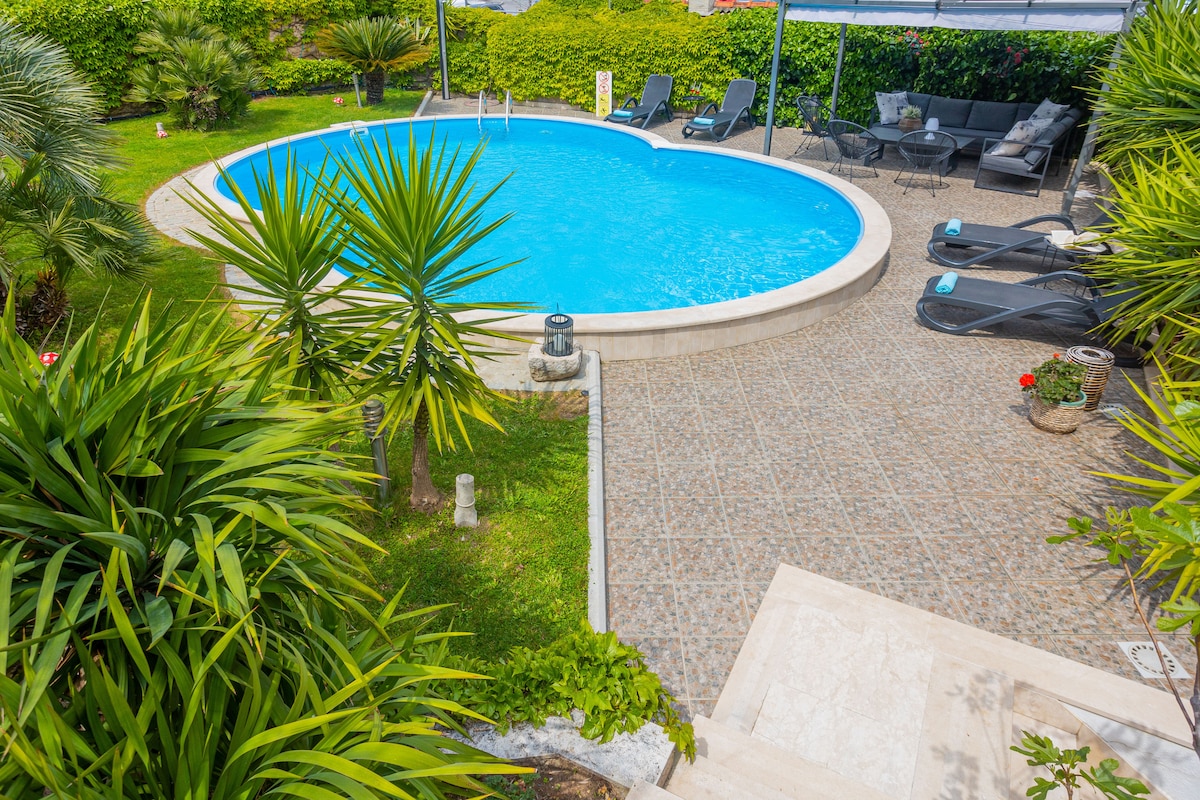 A private pool is surrounded by lush greenery and tropical plants, creating a serene atmosphere. Comfortable lounge chairs are arranged along the pool deck, inviting relaxation. The pool features a rounded shape with clear blue water, reflecting the ample sunlight in the outdoor setting.