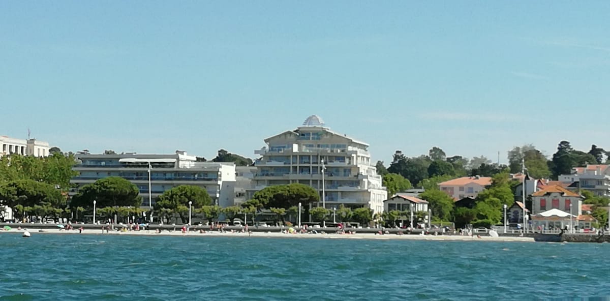 A modern residential building, designed with multiple levels, is situated along the waterfront. Lush trees line the promenade in front, while the sandy beach and calm waters are visible below. Brightly colored buildings create a vibrant backdrop on the distant shoreline.