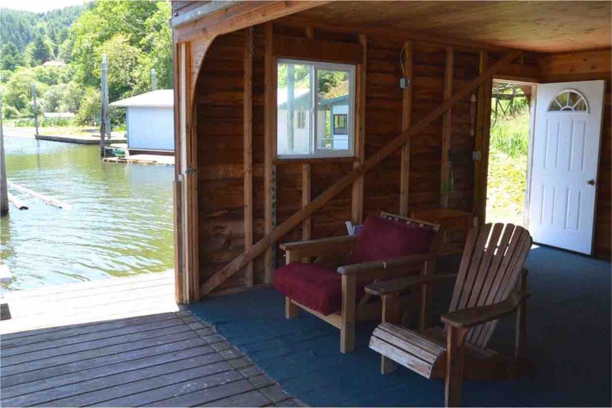A cozy seating area features two wooden chairs, one with cushions, positioned near an open doorway overlooking the water. Natural wooden walls create a warm environment, and a window allows light to brighten the space.